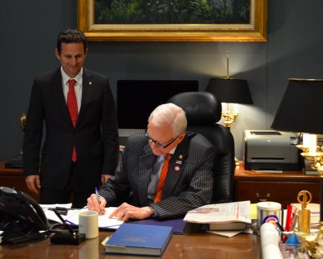 Two men in an office, one signing a bill and the other standing behind him