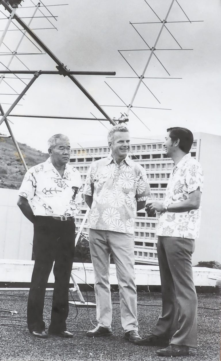 Three men in aloha shirts standing by a satellite