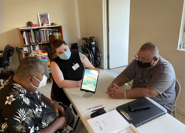 Two men at a desk and a woman wearing a mask helping them to use an ipad