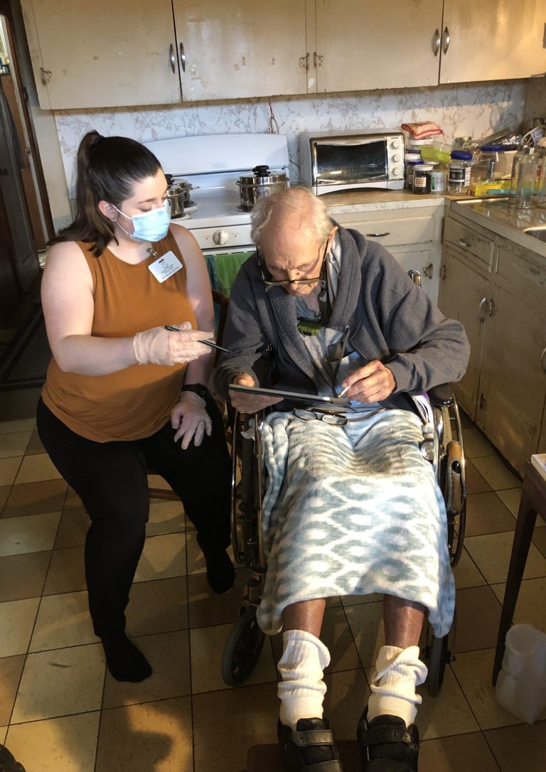 Woman and elderly man in wheelchair in kitchen with the man using a tablet