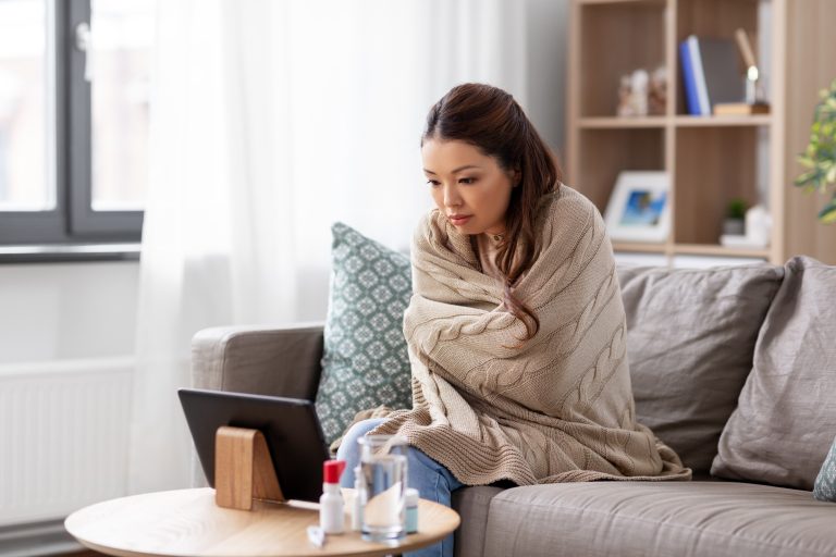 Woman wrapped in blanket on couch next to a table covered in medicine and looking in to a computer screen